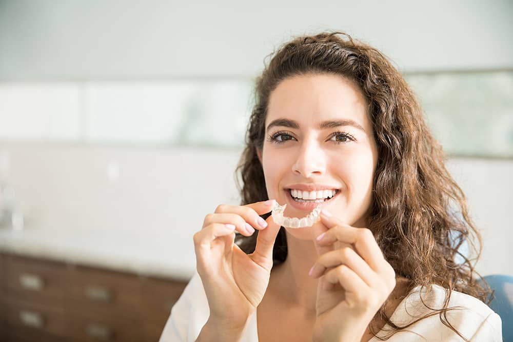 invisalign A woman smiles brightly, gently holding her hand to her mouth, showcasing her teeth.