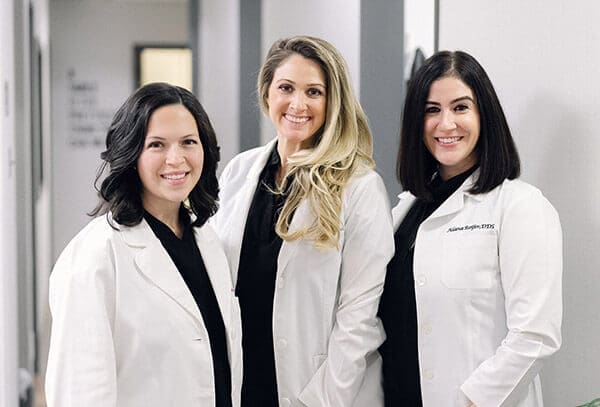 Three women at CR Dental Group stand together in white lab in a hallway, smiling at the camera.